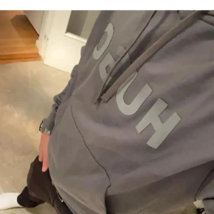 Hugo Boss hoodie  - Säljer en stilren grå hoodie från Hugo med stor logga framtill. Tröjan har huva med snörning, magficka och ribbade muddar. Perfekt för dig som gillar en clean och avslappnad look. Materialet är mjuk bomullsmix som känns skönt mot huden. Storlek M men passar S