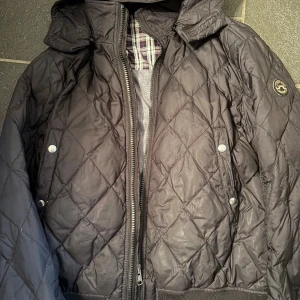 Svart quiltad jacka från J.Lindeberg! - Quiltad svart jacka från J.Lindeberg med huva och dragkedja framtill. Jackan har ett snyggt rutmönster, två fickor med tryckknappar och ett broderat märke på ärmen. Fodret har rutigt tyg och jackan är perfekt för kyliga dagar. Fraktas samma dag och pris kan diskuteras vid snabb affär! (Passar som en M/S) 