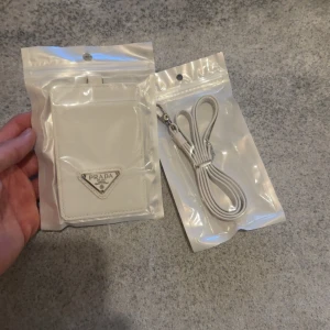 Prada korthållare runt om nacken   - Snygg liten vit axelväska från Prada med klassisk trekantig metall-logga framtill. Väskan har en stilren, rektangulär form och kommer med matchande axelrem. Perfekt för att bära det viktigaste och ge din outfit en lyxig touch.