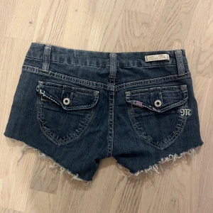 Miss Me jeansshorts med rå kant - Snygga mörkblå jeansshorts från Miss Me med råa, fransiga kanter och dekorativa bakfickor med knapp. Shortsen har låg midja, klassiska bälteshällor och coolt mönstrat tyg på insidan av linningen. Perfekta för sommaren!