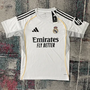 Real Madrid vit matchtröja Adidas - Real Madrid fotbollströja i vitt från Adidas med svarta detaljer och gula linjer. Klassisk klubbmärke på bröstet, Adidas-logga och tre svarta ränder på axlarna. Tillverkad i lätt och ventilerande polyester, perfekt för match eller träning.