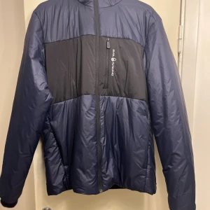 Blå dunjacka från Sail Racing - Snygg dunjacka från Sail Racing i mörkblå och svart färg. Jackan har dragkedja framtill, praktiska fickor med dragkedja och en bröstficka med Sail Racing-logga. Materialet är lätt och syntetiskt, perfekt för kyliga dagar. Passformen är normal och jackan har långärmade ärmar.