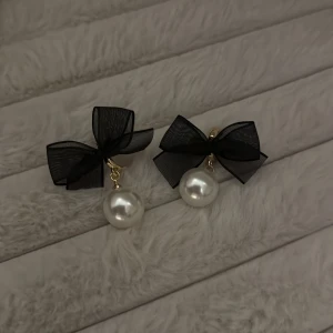 Örhängen med svarta rosetter och pärlor - Supersöta örhängen med stora vita pärlor och svarta rosetter i organza. Fästet är i guldton och designen är både lekfull och elegant. Perfekta för att ge din outfit en extra touch av stil.
