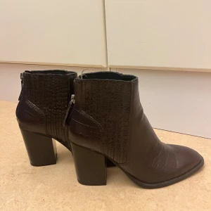 Bruna stövletter med klack från Zara - Snygga mörkbruna stövletter från Zara i croco-mönstrat skinnimitation. Skorna har spetsig tå, hög blockklack och dragkedja bak för enkel påtagning. Perfekta för dig som vill ha en edgy och trendig look.