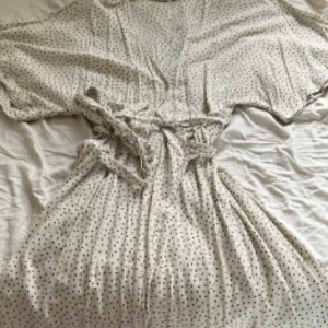 Prickig omlottklänning med knytband - Superfin ljus omlottklänning med små svarta prickar och knytband i midjan. Klänningen har korta, vida ärmar och v-ringning. Perfekt för varma dagar och ger en avslappnad men ändå söt look.