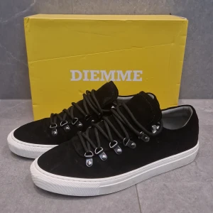 Svarta mockasneakers från Diemme - Snygga svarta sneakers från Diemme i mocka med vit platt sula. Skorna har unika metallöglor och kraftiga svarta snören som ger en cool outdoor-vibe. Perfekta för dig som gillar stilrena men ändå sportiga skor med premiumkänsla.