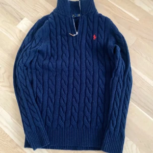 Marinblå stickad tröja Polo Ralph Lauren - Snygg marinblå stickad tröja från Polo Ralph Lauren med klassiskt kabelmönster och röd broderad logga på bröstet. Tröjan har half zip, hög krage och långa ärmar. Perfekt för dig som gillar stilrena och tidlösa plagg.