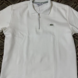 Vit pikétröja med dragkedja från Lacoste - Snygg vit pikétröja från Lacoste med klassisk krage och halv dragkedja framtill. Tröjan har lång ärm och den ikoniska gröna krokodilloggan på bröstet. Perfekt för en stilren och sportig look.