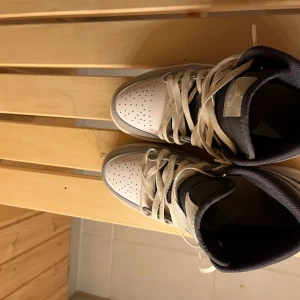 Nike Jordan 1 Mid i grå och vit - Nike Jordan 1 Mid sneakers med snygg grå och vit färgkombination, svart swoosh och klassisk snörning. Skorna har perforerad tå, rund tå och platt sula. Ovandelen är i skinn och syntet, med bekväm vadderad krage i mörkgrått. Perfekt för dig som gillar streetstyle.