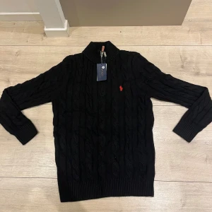 Svart stickad tröja Polo Ralph Lauren - Svart kabelstickad tröja från Polo Ralph Lauren med halv dragkedja och hög krage. Klassisk röd logga broderad på bröstet. Tröjan är långärmad och har ribbade muddar och nederkant. Perfekt för dig som gillar stilren och tidlös design.