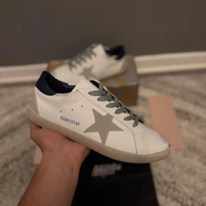 Golden Goose skor  - Golden Goose sneakers i vitt skinn med grå mockadetalj i form av en stjärna på sidan. Skon har grå snörning, beige platt sula och mörkblå häl. Snygg och stilren design med GGDB/SSTAR-tryck på sidan. Perfekt för dig som gillar exklusiva och trendiga sneakers.