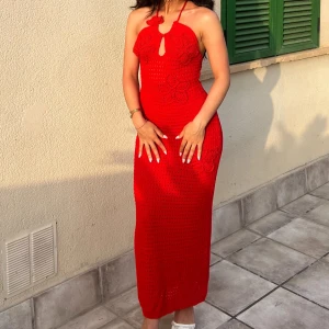 Röd virkad långklänning med blommor - Snygg röd virkad långklänning med halterneck och blommiga detaljer. Klänningen är ärmlös och har en liten öppning framtill vid halsen. Perfekt för varma dagar och ger en trendig boho-vibe.