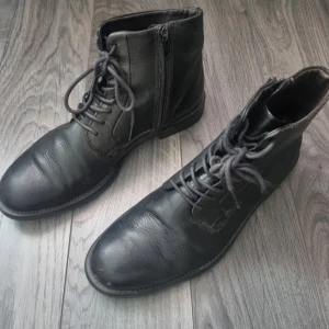 Svarta Vagabond-boots i läder - Svarta kängor från Vagabond i äkta skinn med snörning och dragkedja på sidan.        Man kan se lite att den har varit använt tidigare. 