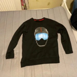 Svart tröja med hjälmtryck från Prada - Cool svart tröja från Prada i storlek L med ett stort tryck av en hjälm och blåa skidglasögon på bröstet. Tröjan har rund halsringning och långa ärmar. Perfekt för dig som gillar unika designer och streetwear vibes.