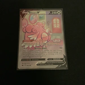 Blissey V Pokémonkort - Blissey V holografiskt Pokémonkort i mycket fint skick, skyddat i plastficka. Kortet har 250 HP och unika attacker som Natural Cure och Blissful Blast. Perfekt för samlare eller spelare som vill stärka sin lek med ett kraftfullt V-kort.