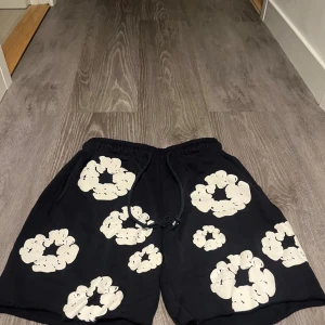Svarta Denim Tears shorts med blommor - Svarta shorts från Denim Tears med vita blommiga tryck över hela plagget. De har elastisk midja med snörning, råa kanter nertill och en bakficka. Perfekta för en chill sommarlook och riktigt sköna att bära.
