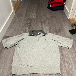 Grå half zip tröja Polo Ralph Lauren - Snygg grå tröja från Polo Ralph Lauren med mörkare grå krage och halv dragkedja. Klassisk broderad logga på bröstet och kortärmad modell. Perfekt för en chill och stilren look.