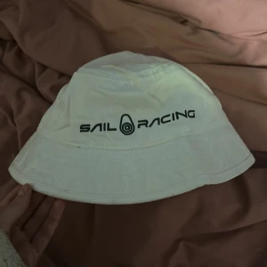 Vit Sail Racing bucket hat - Vit bucket hat från Sail Racing med svart logga broderad framtill. Hatten är tillverkad i 100% bomull och har klassisk form med bred brätte. Perfekt för soliga dagar och en clean streetstyle-look.