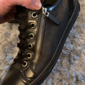 Svarta Louis Vuitton sneakers med dragkedja - Säljer ett par svarta Louis Vuitton sneakers i skinn med klassiskt rutmönster och cool dragkedjedetalj på sidan. Skorna har snörning, rund tå och platt sula. Perfekta för dig som gillar lyxiga och stilrena detaljer.