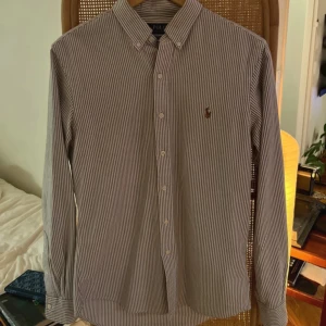 Ralph lauren skjorta  - Klassisk randig skjorta från Polo Ralph Lauren i slim fit. Skjortan är beige/vit med tunna ränder, har knappar framtill och lång ärm. Broderad logga på bröstet. Perfekt för en clean och stilren look.