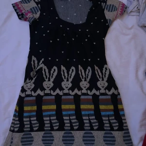 Mönstrad kortklänning från Smash! - Unik kortklänning från Smash! med coolt kaninmönster, små hjärtan och färgglada ränder. Klänningen har korta ärmar med grafiskt mönster och knytband i midjan. Färgerna är svart, grå, blå, röd, gul och beige. Perfekt för dig som gillar lekfull stil och detaljer.