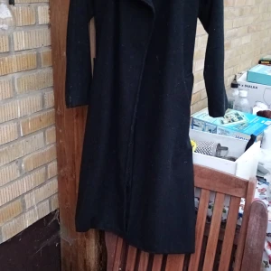 Svart lång ullkappa med bälte stl M - Säljer en svart lång ullkappa med bälte i midjan och klassisk krage. Jackan har långa ärmar och är tillverkad av en blandning av polyester, ull, viskos och akryl. Perfekt för dig som gillar stilrena och tidlösa plagg.stl M
