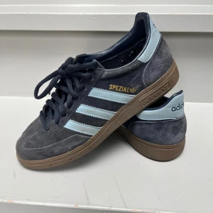 Adidas Spezial sneakers mörkgrå/blå - Säljer ett par Adidas Spezial sneakers i mörkgrå mocka med ljusblå detaljer och klassiska tre ränder på sidorna. Skorna har bruna gummisulor, snörning och guldfärgad Spezial-logga. Perfekta för dig som gillar retrostil och streetwear.