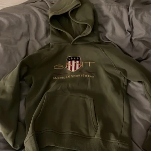 Olivgrön hoodie från GANT - Snygg olivgrön hoodie från GANT med broderad logga och sköld på bröstet. Klassisk känguruficka framtill och huva. Mjuk insida och ribbade muddar. Perfekt för dig som gillar sportig och avslappnad stil.