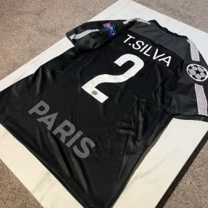 PSG T. Silva svart matchtröja Nike M - Säljer en svart Paris Saint-Germain fotbollströja från Nike med T. Silva och nummer 2 på ryggen. Tröjan har korta ärmar, Dri-FIT-material och detaljer i vitt, inklusive klubbmärke, sponsorlogga och Champions League-patch på ärmen. Perfekt för dig som älskar PSG.
