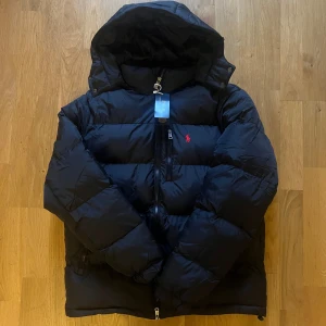 Svart pufferjacka från Polo Ralph Lauren - Svart pufferjacka från Polo Ralph Lauren med huva och tvåvägsdragkedja. Perfekt för kalla dagar och har en modern, loose passform| skriv vid funderingar                                                                Priset kan självklart diskuteras!!