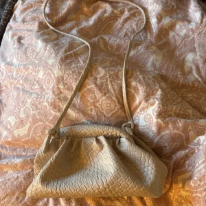 Beige axelväska i flätat skinn - Snygg beige axelväska med lång rem och flätad design. Väskan har en mjuk, slouchy form och är tillverkad i skinn med ett invändigt foder. Perfekt för dig som gillar stilrena och trendiga accessoarer. Lite smutsig inuti 