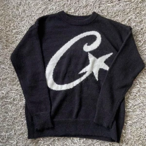 Svart Corteiz med svart mössa  - Svart mössa från Corteiz med stor vit logga och stjärndetaljer framtill. Snyggt stickad i mjukt material som håller dig varm och ger en clean streetwear-look. Perfekt för dig som vill ha en stilren accessoar med tydlig branding.