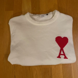 Vit stickad tröja från Ami Paris - Säljer en vit stickad tröja från Ami Paris med röd hjärtlogga och A på bröstet. Tröjan har rund halsringning, långa ärmar och bred ribbad mudd nedtill. Perfekt för dig som gillar stilrena plagg med en twist.