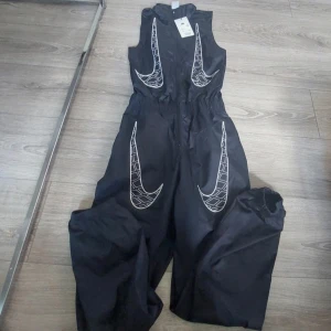 Nike svart jumpsuit med vita detaljer XS + byxor  - Nike svart jumpsuit i syntetmaterial med stora vita Swoosh-loggor framtill och baktill. Ärmlös överdel, resår i midjan och dragkedja framtill. Byxorna har elastiska benslut för en sportig vibe. Perfekt för dig som gillar streetwear och vill sticka ut.   Säljes för 800 kr så ett kap 💕