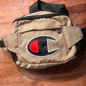 Champion väska - Snygg beige magväska från Champion med stort broderat logotypmärke i svart, vitt och rött framtill. Ca 30cm. Väskan har justerbart svart band, dragkedja och praktisk ficka på framsidan. Rymliga fack..