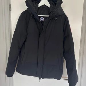 Svart dunjacka från Canada Goose M - Svart dunjacka från Canada Goose med huva och snygga detaljer som snörning vid kragen. Jackan har två sidofickor och klassisk logga på ärmen. Perfekt för kalla dagar och har en clean, mörk look som funkar till allt.