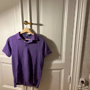 Lila pikétröja från Polo Ralph Lauren - Säljer en klassisk lila pikétröja från Polo Ralph Lauren med grön broderad logga på bröstet. Tröjan har korta ärmar, krage och knappar vid halsen. Tillverkad i mjuk bomull som är skön att bära.