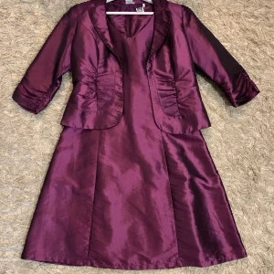 Långklänning med matchande kavaj - Säljer en elegant långklänning i glansigt material med v-ringning och figurnära passform. Till klänningen hör en matchande kavaj med trekvartsärm, snygga rynkdetaljer och klassisk krage. Perfekt för dig som vill sticka ut med en lyxig look.