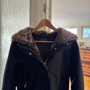 Svart parkas med fuskpäls från Jofama - Säljer en svart parkas från Jofama med snygg fuskpäls runt huvan. Jackan har långa ärmar, dragkedja framtill och flera fickor med både dragkedja och knappar. Perfekt för dig som vill ha en varm och stilren look under kalla dagar. Om du har några funderingar är det bara att höra av sig🫡