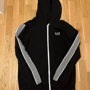 Svart EA7 hoodie med vita detaljer - Svart hoodie från EA7 Emporio Armani med vita ränder längs ärmarna och vita dragkedjor. Tröjan har huva, lång ärm och EA7-logga på bröstet. Perfekt för dig som gillar sportig och stilren streetwear.