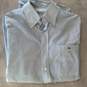 Blårandig skjorta från Lacoste storl. 39 - Snygg skjorta från Lacoste med tunna blå och vita ränder, klassisk krage och knappar framtill. På bröstfickan sitter den ikoniska gröna krokodilen broderad. Skjortan är långärmad och gjord i mjuk bomull som känns skön mot huden.