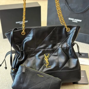 Svart quiltad axelväska från Saint Laurent - Snygg svart axelväska från Saint Laurent i quiltat skinn med guldiga kedjehandtag och ikoniskt YSL-emblem framtill. Väskan har dragsko i sidorna och medföljande matchande clutch. Perfekt för dig som vill ha en lyxig och trendig accessoar.