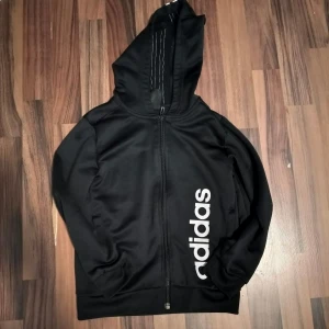 Svart Adidas hoodie med dragkedja - Svart hoodie från Adidas med hel dragkedja och stor vit logga längs sidan. Klassiska tre vita ränder på huvan. Tillverkad i mjuk polyester, perfekt för träning eller chill. Långärmad modell med huva och ribbade muddar. Barn storlek 128