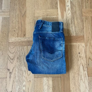 Replay Anbass blå jeans - Snygga Replay Anbass jeans i klassisk blå tvätt med slitningar och ljusare partier. Modellen har fem fickor, normal midja och raka ben. Jeansen är tillverkade i mjukt denimtyg och har Replay-logga på bakfickan och läderpatch i midjan. Strl, 32,32