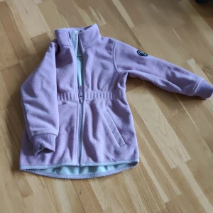 Ljuslila fleecejacka Polarn O. Pyret - Säljer en ljuslila fleecejacka från Polarn O. Pyret med hög krage, hel dragkedja och ribbade muddar. Jackan har ett broderat märke på ärmen och praktiska fickor framtill. Perfekt för kyliga dagar och enkel att matcha med andra plagg.