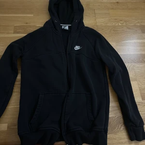 Svart Nike hoodie med dragkedja (barn storlek) - Svart hoodie från Nike med vit logga på bröstet. Tröjan saknar dragsnören till huvan , två fickor framtill och hel dragkedja. Perfekt för chill eller när du vill ha en enkel streetstyle-look.