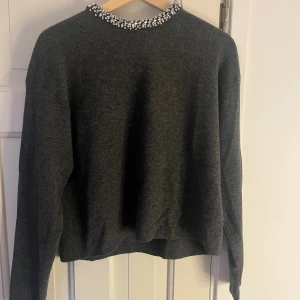 Mörkgrå stickad tröja med glittriga prälor/stenar  - Supersnygg mörkgrå stickad tröja med lång ärm och croppad passform. Runt halsen sitter glittriga pärlor/stenar. Den är super skön och har ett mjukt kvalite, men säljer då den inte kommer till användning☺️😊