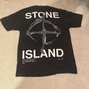 Svart Stone Island t-shirt med tryck - Svart t-shirt från Stone Island med stort grafiskt tryck och logga på ryggen samt liten logga på bröstet. Klassisk passform och rund hals. Materialet är mjuk bomull som känns skön mot huden. Perfekt för dig som gillar streetwear och vill sticka ut.