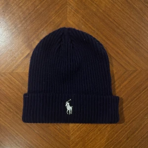 Mörkblå Ralph Lauren mössa - Mörkblå ribbstickad mössa från Polo Ralph Lauren med klassisk broderad logga framtill. Tillverkad i mjuk ull som håller dig varm under kyliga dagar. Enkel och stilren design med uppvikt kant.
