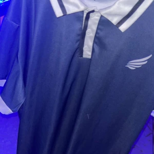 Blå fotbollströja med vit krage - Snygg blå fotbollströja med vit krage och knappslå. Tröjan har korta ärmar med vita detaljer och ett stilrent tryck av en vinge på bröstet. Materialet är lätt och andas, perfekt för träning eller match. Klassisk design med sportig känsla.